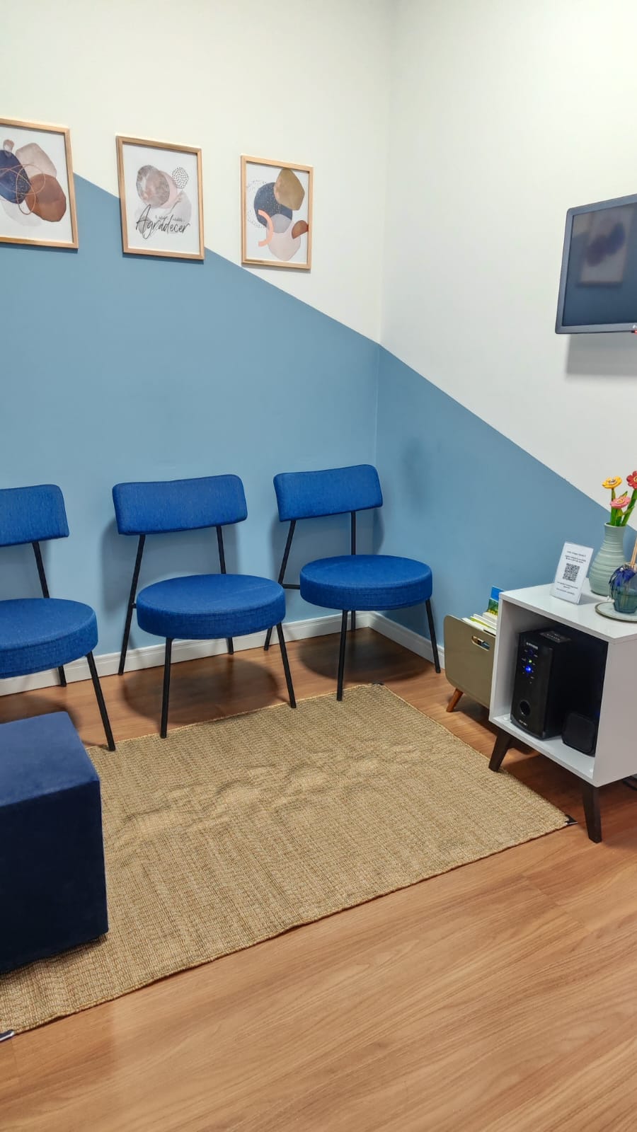 Sala de espera com poltronas azuis, quadros e TV